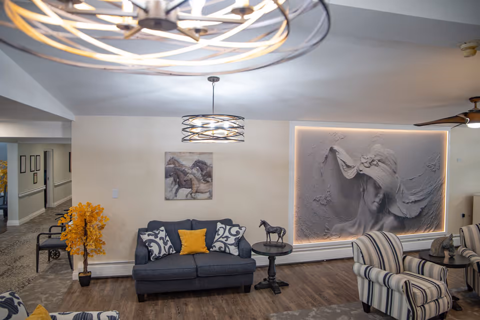 A cozy senior living facility common area with a dark gray sofa adorned with patterned and yellow pillows, a small round side table with a horse figurine, and two striped armchairs with another small table between them. The room features a large textured wall art of a woman wearing a wide-brimmed hat, illuminated by soft lighting. A modern circular ceiling light fixture and a ceiling fan are visible, along with a hallway leading to other rooms. There is also a decorative yellow plant near the sofa.