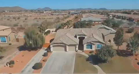 Aerial view of a single-story residential building with a tiled roof, a driveway leading to a two-car garage, and surrounding desert landscaping with sparse vegetation and other houses in the background.