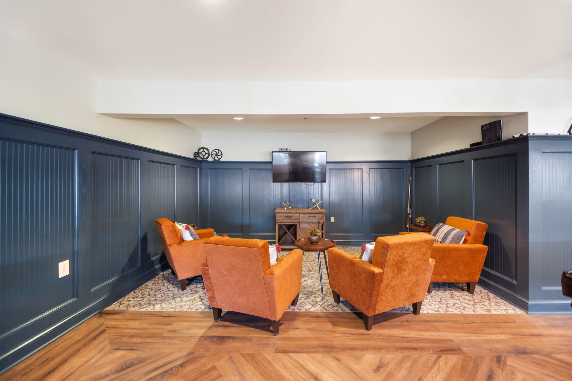 Cozy seating area with four orange armchairs arranged around a small table facing a wall-mounted TV in a wood-paneled room.