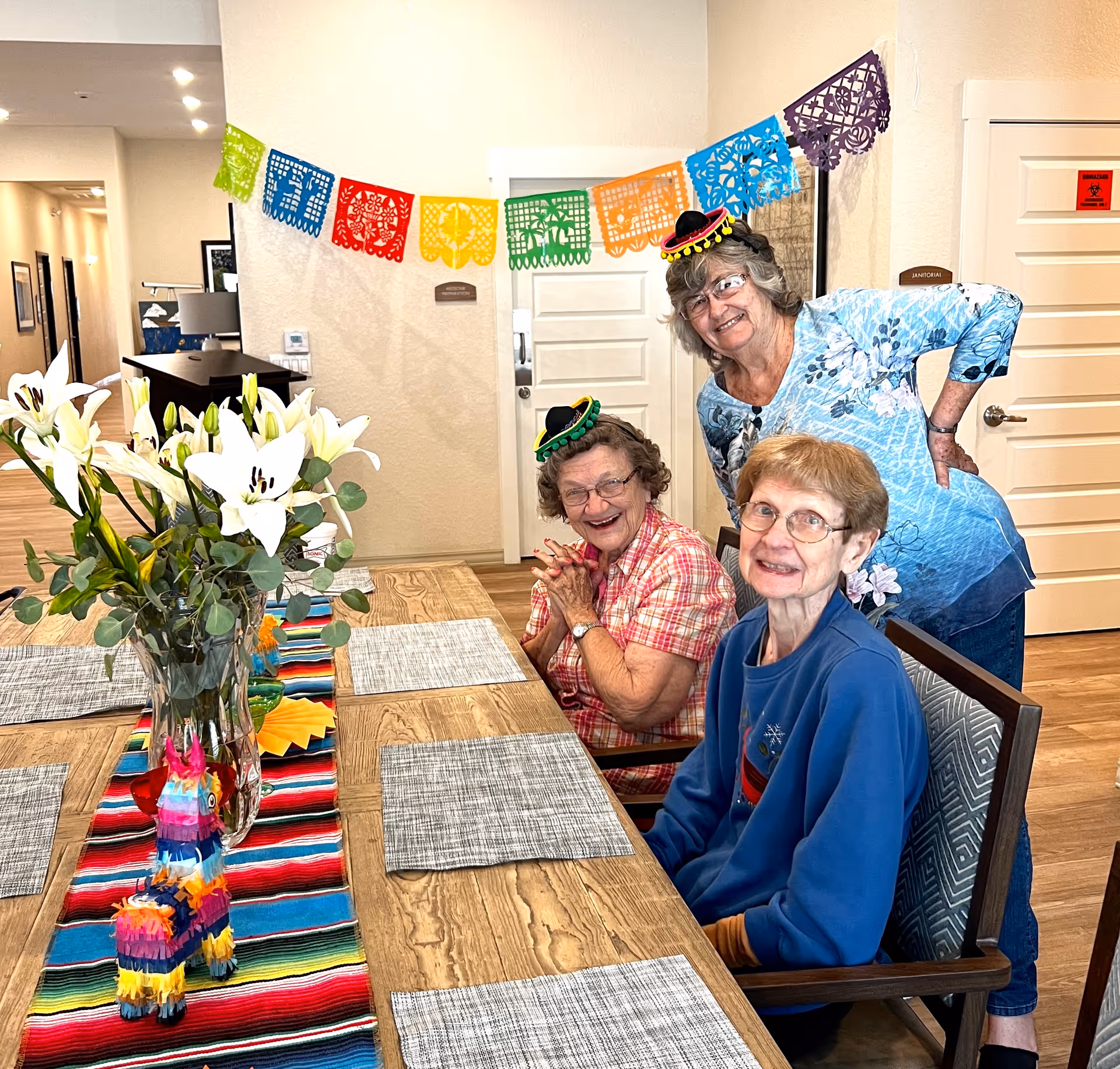 Three elderly women smiling and posing at a wooden dining table decorated with a colorful striped table runner, a vase of white lilies, and a small colorful piñata. Two women are seated wearing small festive hats, while the third woman stands behind them. Colorful paper decorations hang on the wall behind them in a well-lit room with light-colored walls and wooden flooring.
