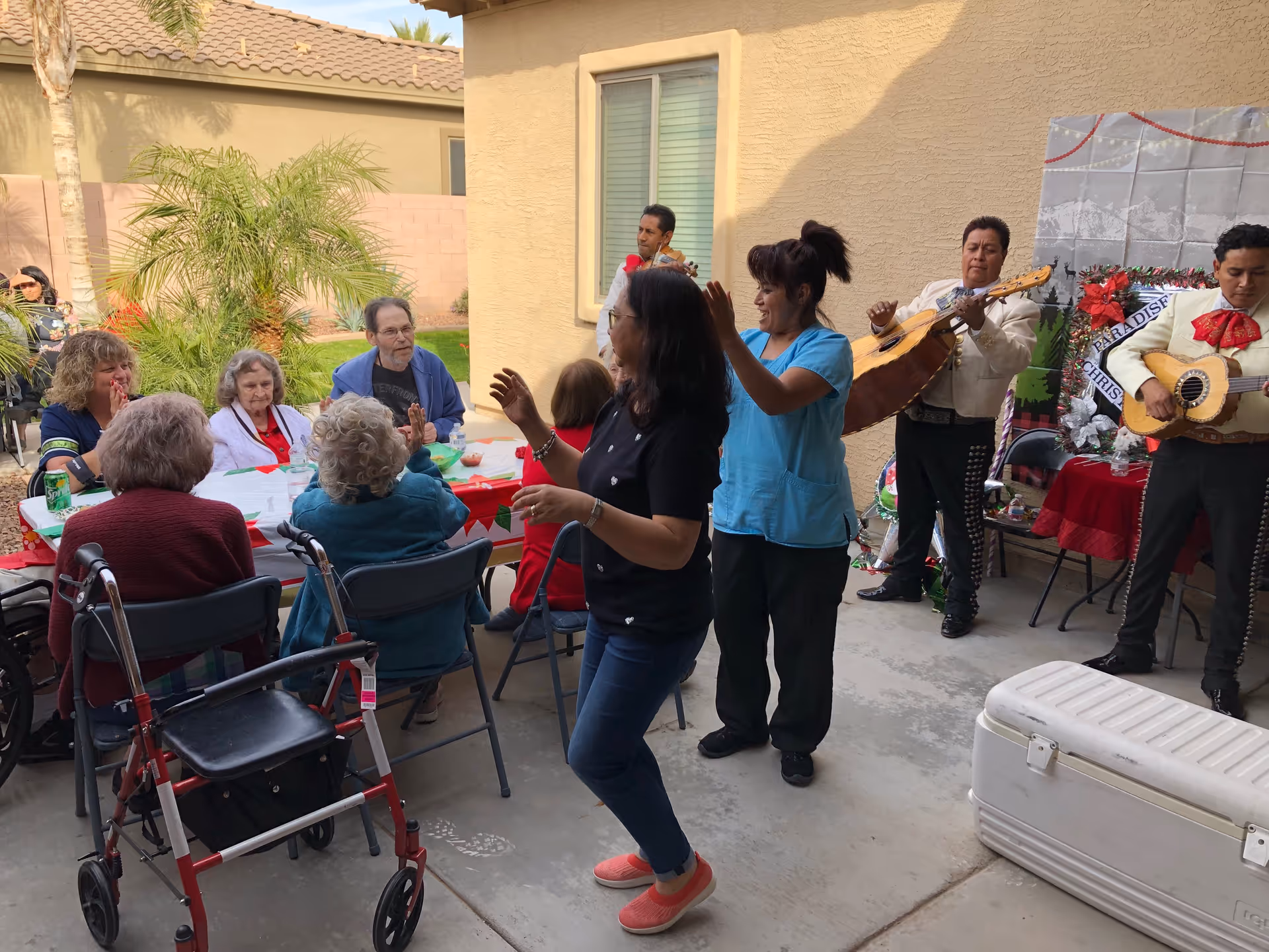 A group of elderly people seated around a table outdoors, enjoying a festive gathering with two women dancing and a mariachi band playing music nearby. The setting is a patio area with palm trees and a beige stucco building wall in the background.