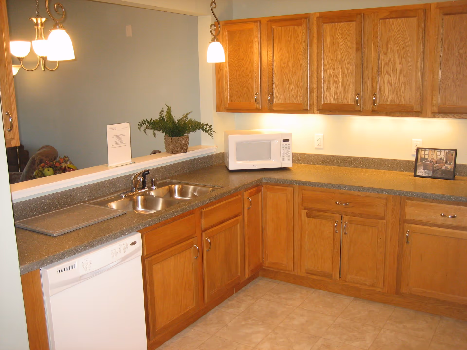 A kitchen area with wooden cabinets, a double stainless steel sink, a white dishwasher, and a white microwave on the countertop. There are two pendant lights hanging above the counter and a small plant and framed picture on the counter. The floor is tiled and the walls are painted light colors.