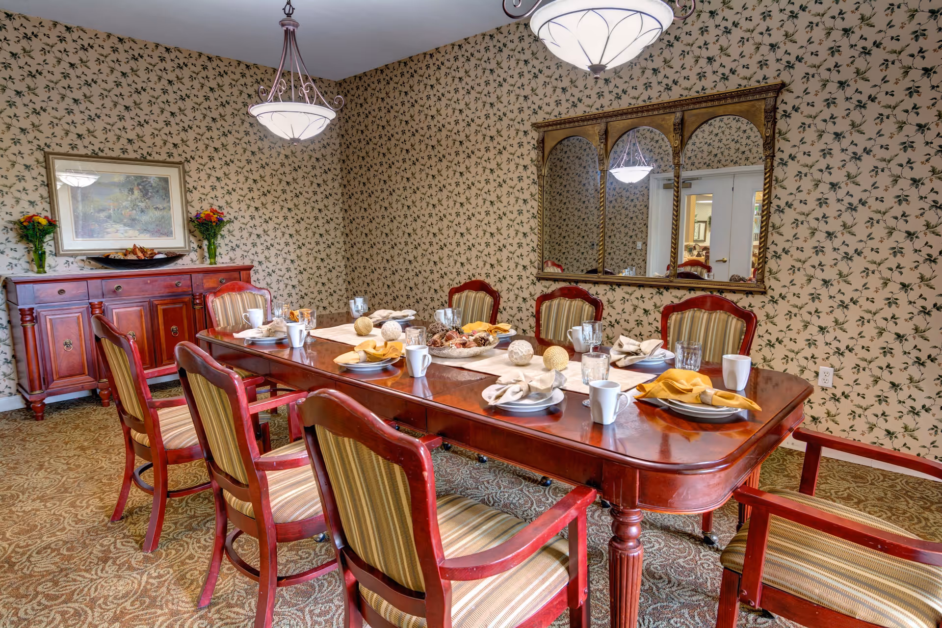 A dining room with a long wooden table set for eight people with plates, napkins, cups, and glasses. The chairs have wooden frames with striped upholstery. The room has floral wallpaper, a large mirror on one wall, a wooden sideboard with flowers and a framed painting above it, and two ceiling light fixtures.