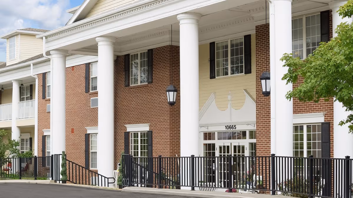 Two-story brick building entrance with large white columns, a covered portico, and a black metal fence.