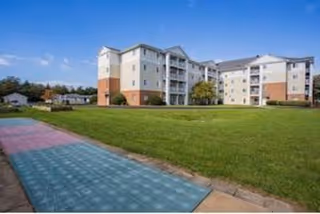 Exterior view of a multi-story senior living facility building with balconies, surrounded by a large green lawn and a colorful shuffleboard court in the foreground under a blue sky with some clouds.