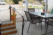 Covered outdoor patio with a glass-top table and chairs beside a small ramp and wooden steps overlooking a gravel driveway.