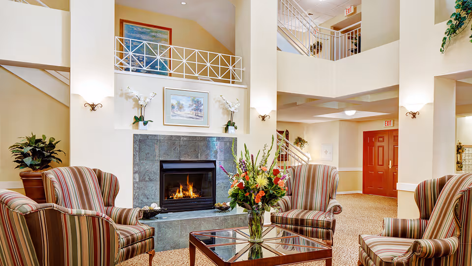 A cozy senior living facility common area with four striped armchairs arranged around a glass coffee table holding a colorful flower arrangement. A lit fireplace with a tiled surround is centered on the far wall, flanked by two white orchid plants and a framed painting above. The room features high ceilings with a second-floor balcony railing visible, warm lighting, and a red exit door in the background.