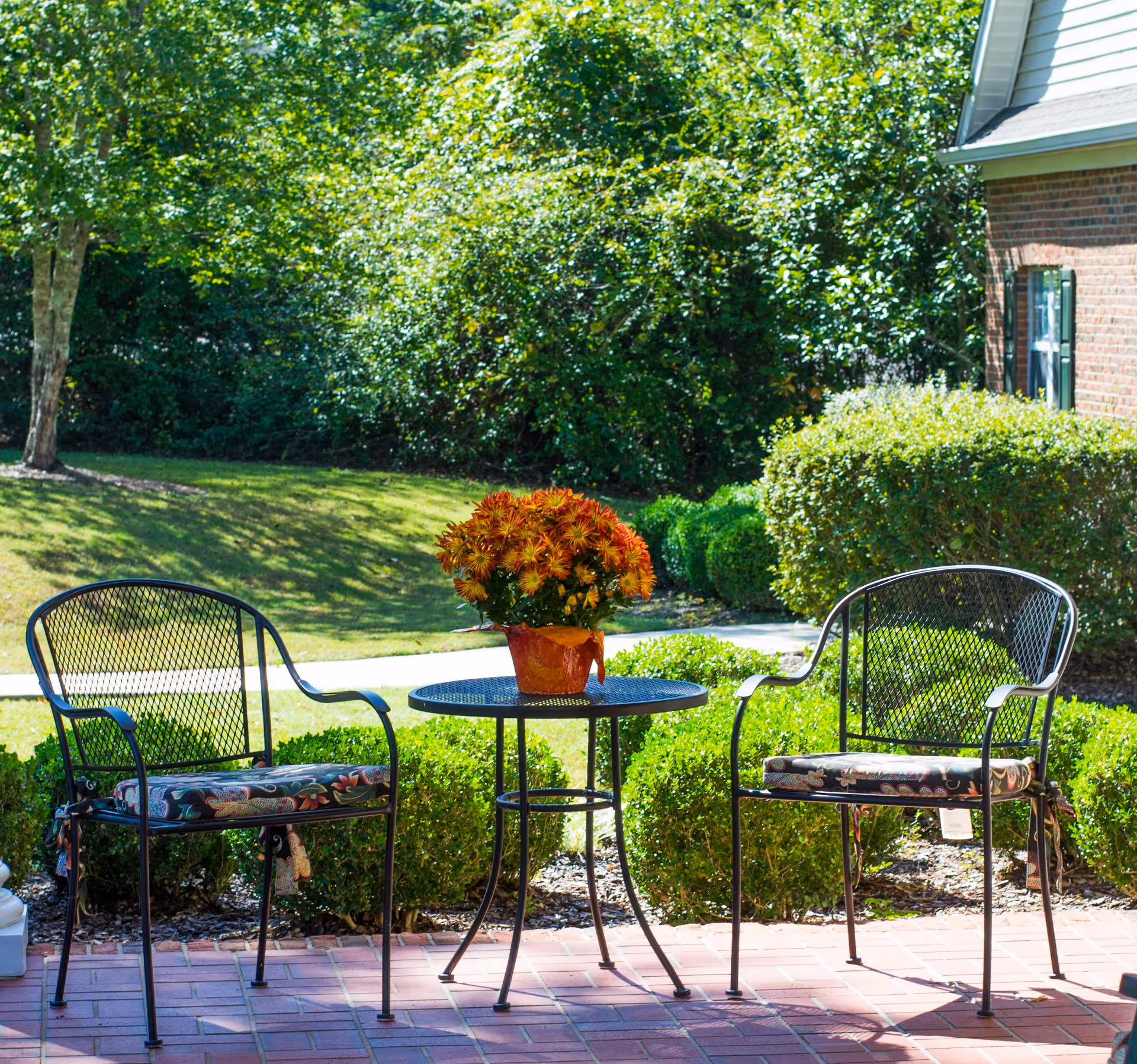 Outdoor patio area with two metal chairs with floral cushions and a small round metal table between them holding a pot of orange flowers. The patio is surrounded by green bushes and trees with a brick building partially visible on the right.