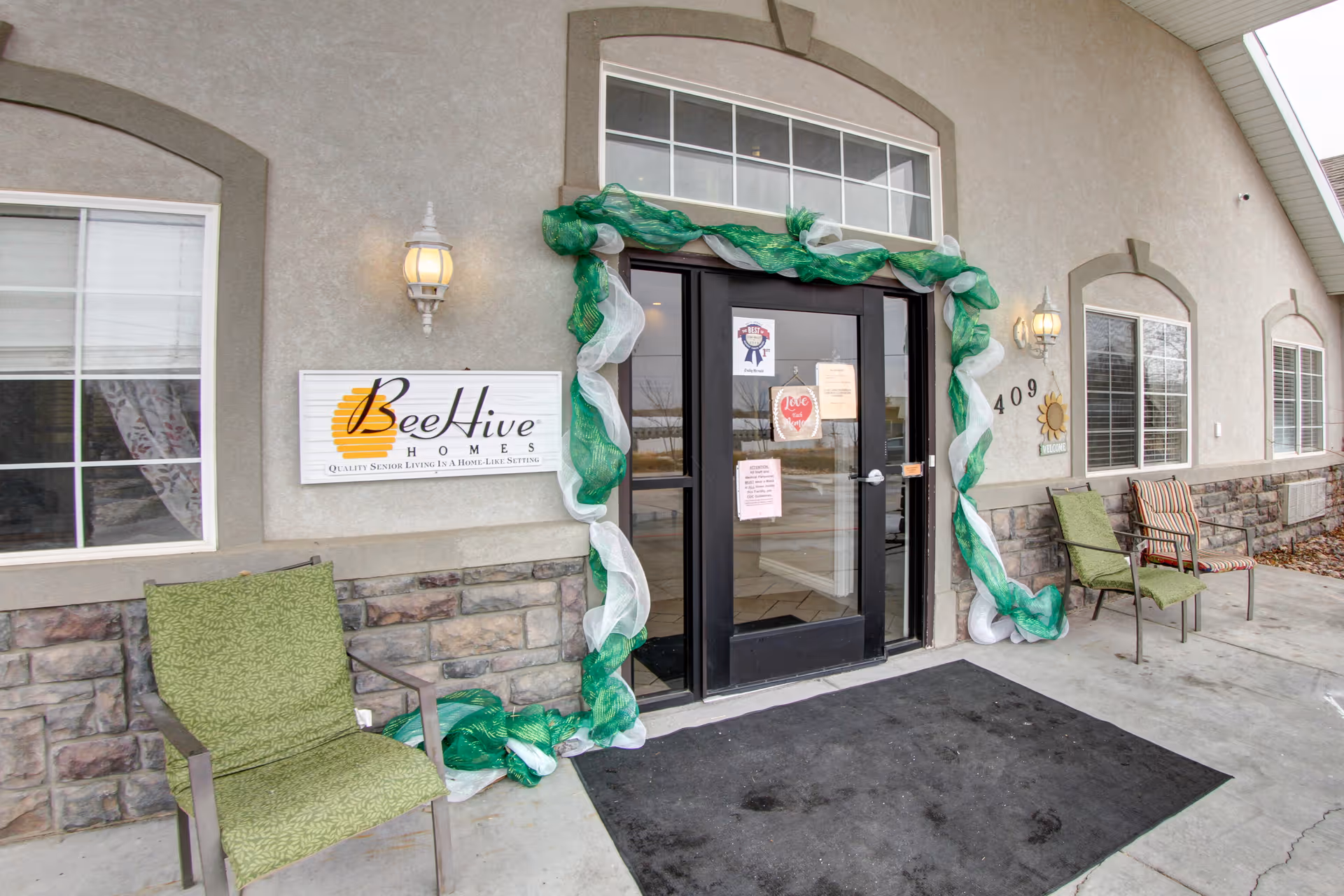Front entrance of Beehive Homes with double glass doors, a Beehive Homes sign, green and white decorative garland, and chairs on the covered porch.