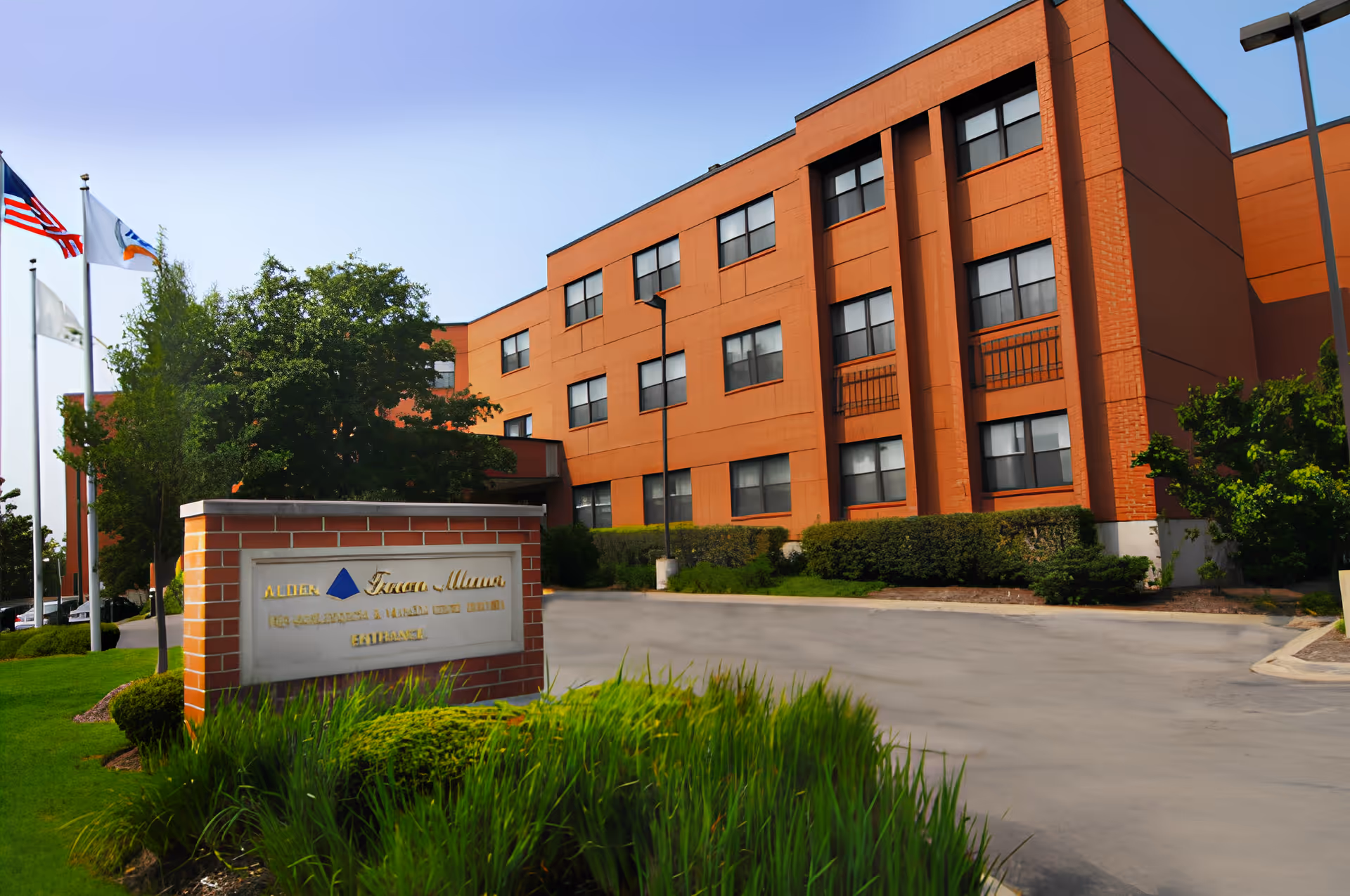 Exterior view of Alden Town Manor Rehab & Health Care Center, a three-story orange brick building with multiple windows. In front of the building is a sign with the facility's name and entrance information, surrounded by green bushes and grass. Several flagpoles with flags are visible on the left side of the image under a clear blue sky.