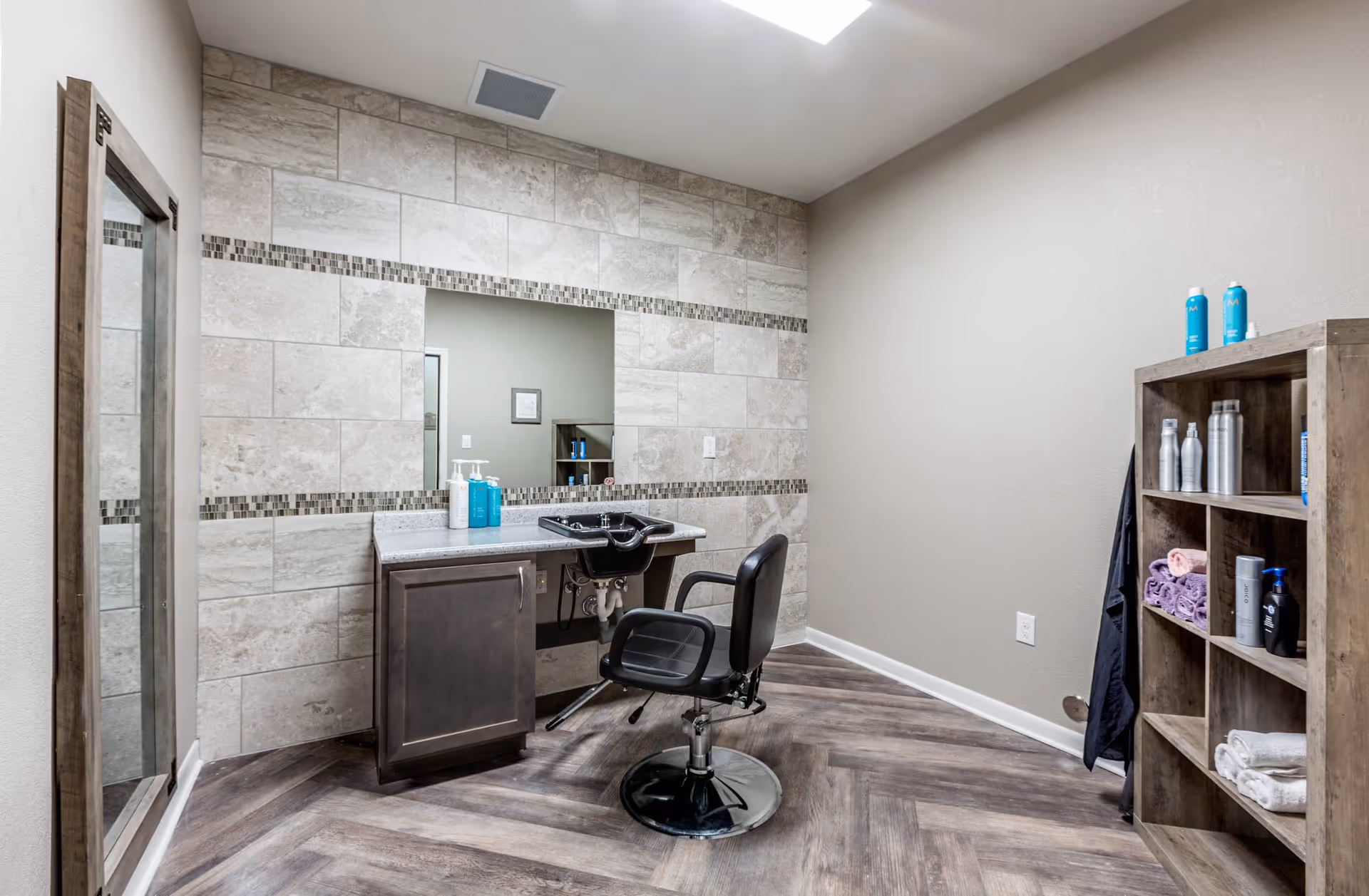 A small salon area with a black salon chair in front of a counter with a built-in sink. The wall behind the counter is tiled with a decorative horizontal strip and has a large rectangular mirror. To the right, there is a wooden shelving unit holding various hair care products and towels. The floor has a wood-patterned design, and the walls are painted light gray.