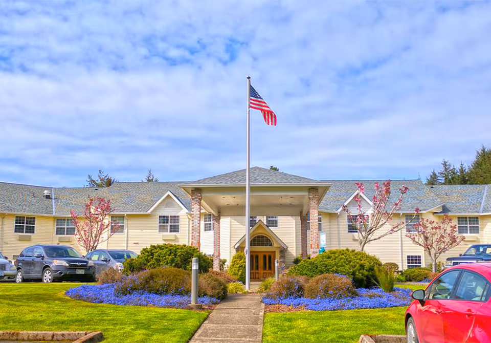 Front entrance of a yellow senior living building with an American flag, landscaped flower beds and parked cars.