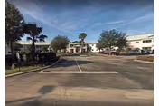 Wide driveway leading to the front entrance of a single-story senior living facility building with palm trees and parked cars on either side under a partly cloudy sky.