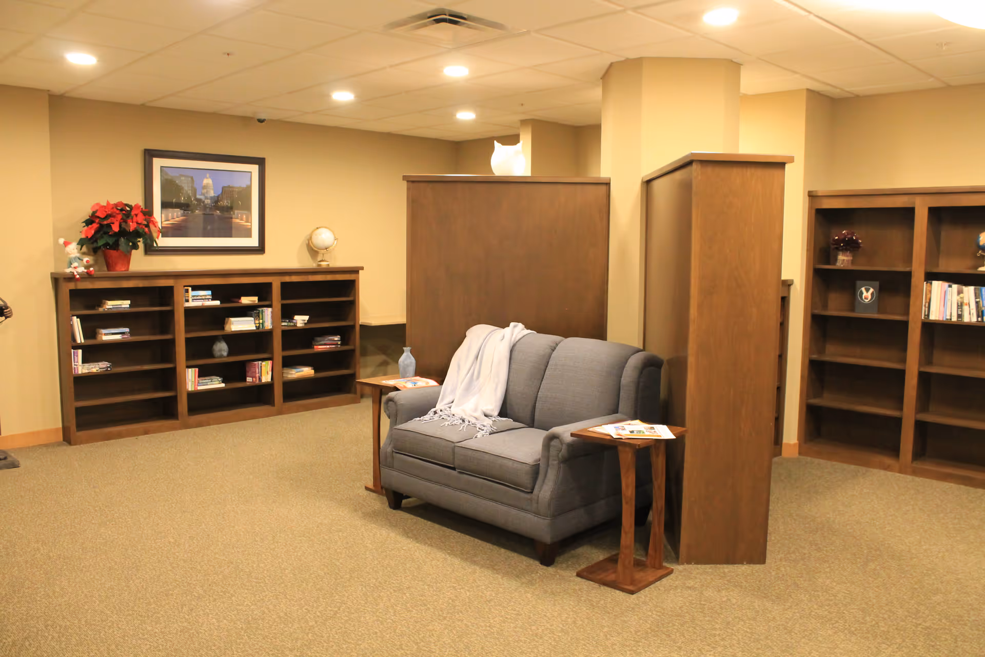A cozy reading nook in a senior living facility featuring a gray loveseat with a white throw blanket draped over it, flanked by two small wooden side tables holding magazines. Surrounding the seating area are wooden bookshelves with various books and decorative items. The walls are beige, and there is a framed picture and a potted plant on one of the shelves.