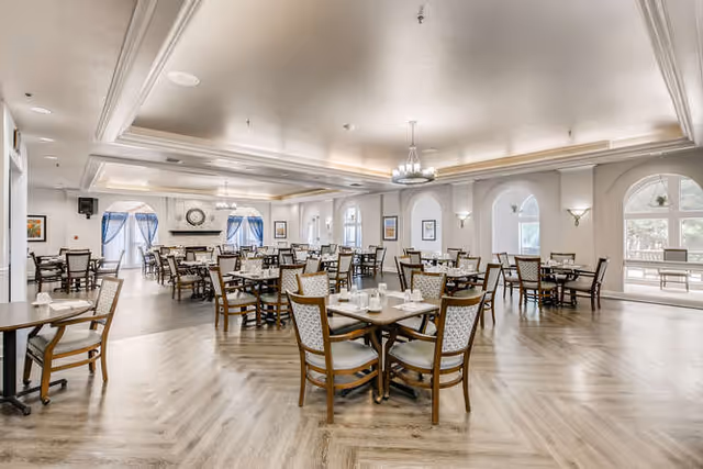 Spacious dining room in a senior living facility with multiple wooden tables and chairs arranged neatly. The room features large arched windows with blue curtains, a decorative ceiling with recessed lighting, and a fireplace with a clock above it. The floor has a light wood pattern, and the walls are painted in a soft neutral color.