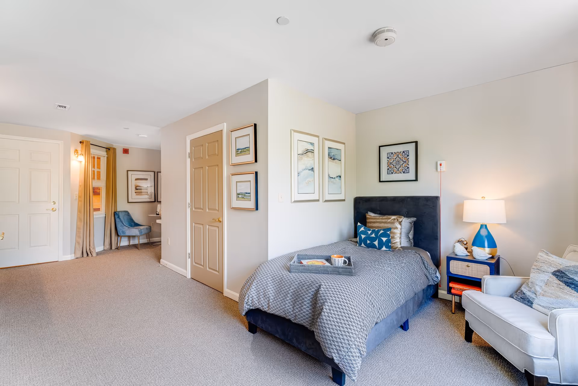 A bright and cozy bedroom in a senior living facility featuring a single bed with gray bedding and decorative pillows, a small side table with a blue and white lamp, a comfortable armchair, and framed artwork on the walls. The room has beige walls and carpeted flooring, with a door and a small seating area with a blue chair near a window with beige curtains.