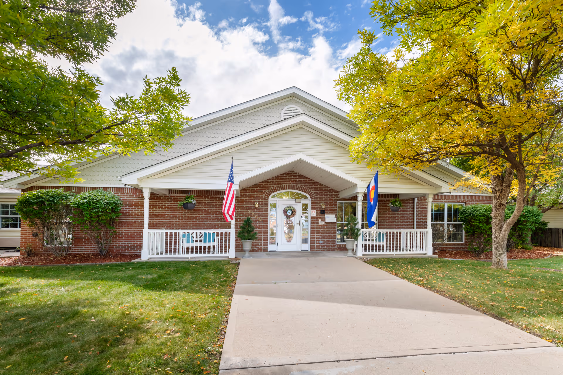 Front exterior view of a single-story brick building with a white porch and two flags, one American and one Colorado state flag, flanking the entrance. The building is surrounded by green grass, bushes, and trees with some yellow leaves under a partly cloudy sky.