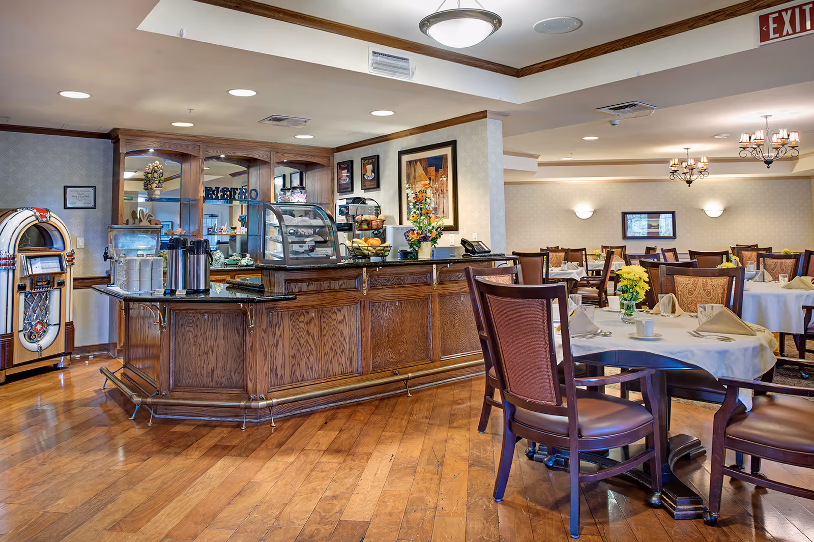 Bright dining room with a wooden service counter, tables set with napkins and flowers, and a jukebox in the corner.