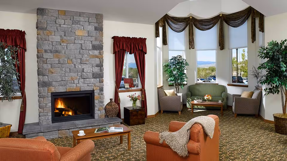 A cozy living room with a stone fireplace burning firewood, surrounded by red curtains on windows. The room has patterned carpet flooring, an orange armchair with a beige throw blanket, a wooden coffee table with a cup and an open book, and a seating area with a green sofa and two gray armchairs near large windows with brown valances. There are also two potted plants in the room.