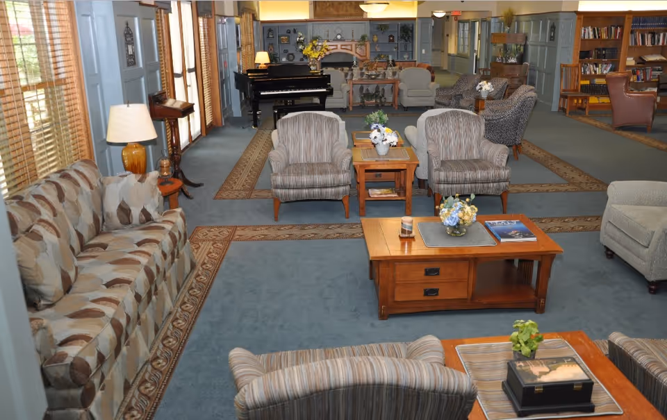 Communal lounge in a senior living facility with sofas and armchairs arranged around coffee tables and a piano in the background.