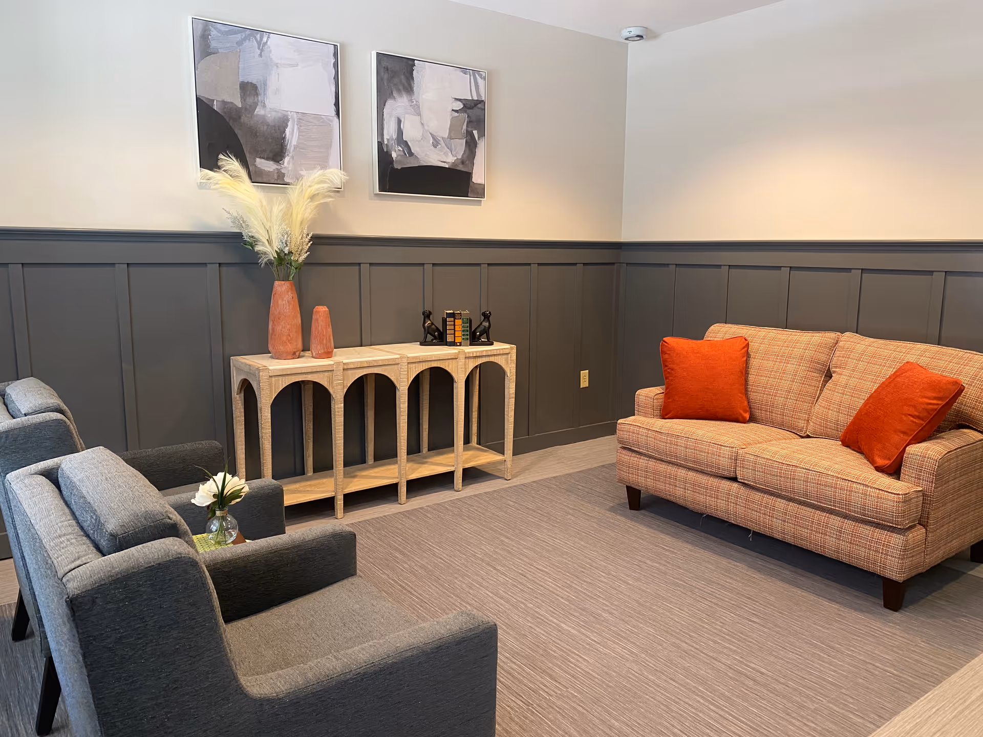 A cozy living room with a plaid sofa adorned with two orange throw pillows, two gray armchairs, a light wood console table with decorative vases and books, and two abstract paintings on the wall above dark gray wainscoting.