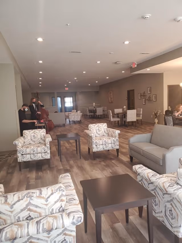 Spacious assisted living common area with couches, patterned armchairs, dining tables, and two masked musicians near the back.