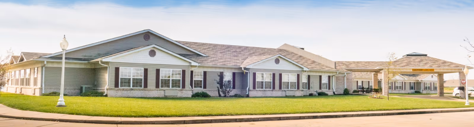 Front exterior of a single-story assisted living building with a covered entrance, green lawn, and street lamps.