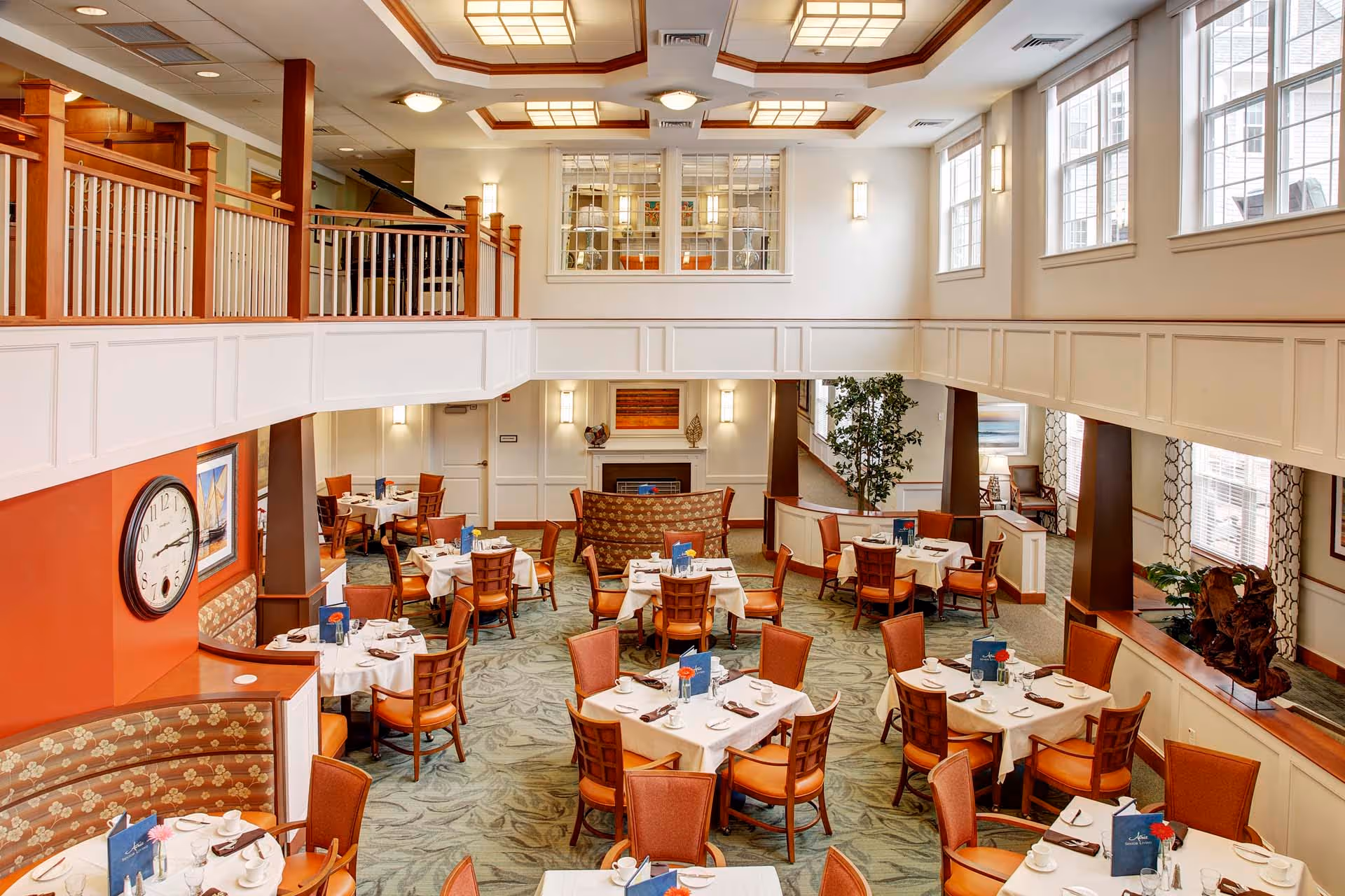 A spacious, well-lit dining room with multiple tables covered in white tablecloths, each set with cups, glasses, napkins, and menus. The room features large windows on the right side, a decorative fireplace at the far end, and a second-floor balcony with wooden railings overlooking the dining area. The walls are painted in warm tones with some artwork and a large clock visible on the left wall.