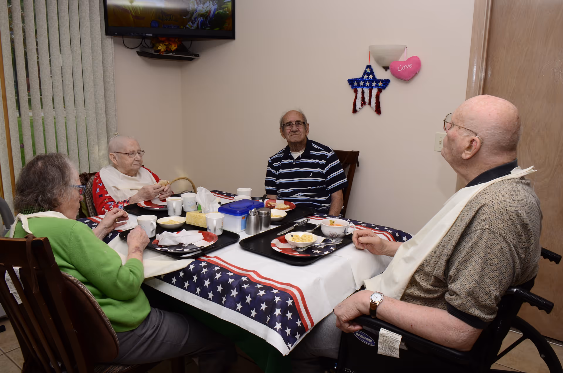 Four elderly individuals sitting around a dining table covered with a patriotic-themed tablecloth featuring stars and stripes. They are having a meal with trays, bowls, cups, and utensils on the table. The room has beige walls with a star decoration and a heart-shaped ornament hanging on the wall. A television is mounted on the wall in the corner.