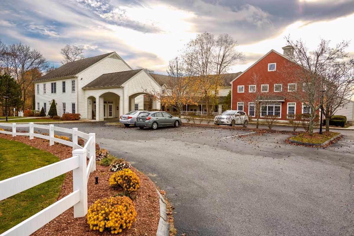 Exterior view of Benchmark at Billerica Crossings facility showing a two-story building with white and red siding, a parking lot with several cars, a white fence, landscaped flower beds with yellow flowers, and trees with autumn foliage under a partly cloudy sky.