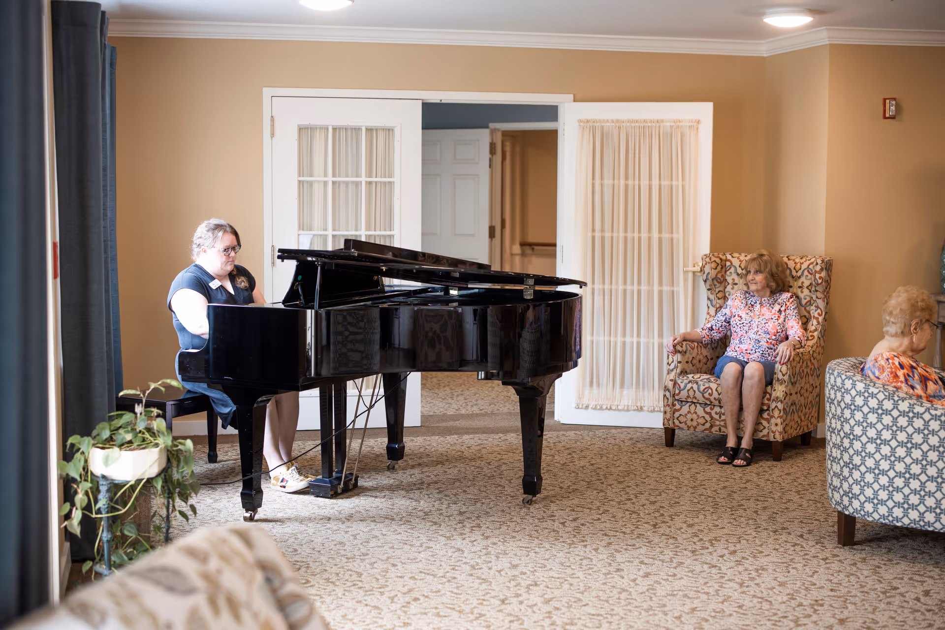 A woman playing a black grand piano in a senior living facility common area while two elderly women sit in patterned armchairs nearby, listening. The room has beige walls, carpeted floor, and white double doors with sheer curtains.
