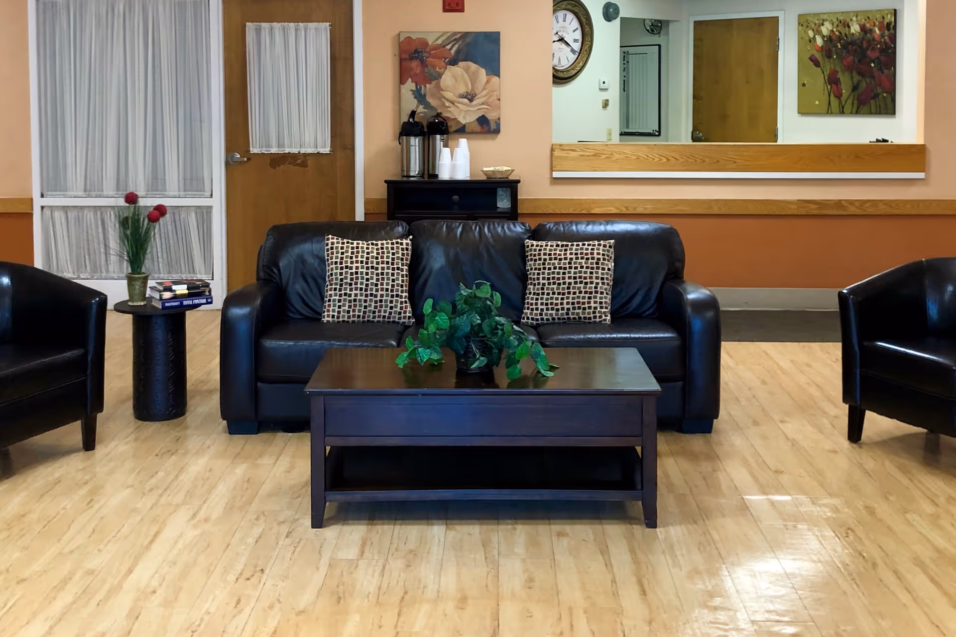 Lobby-style sitting area with a black leather sofa flanked by armchairs, a coffee table with a potted plant, a side table with books and flowers, and artwork on the wall.