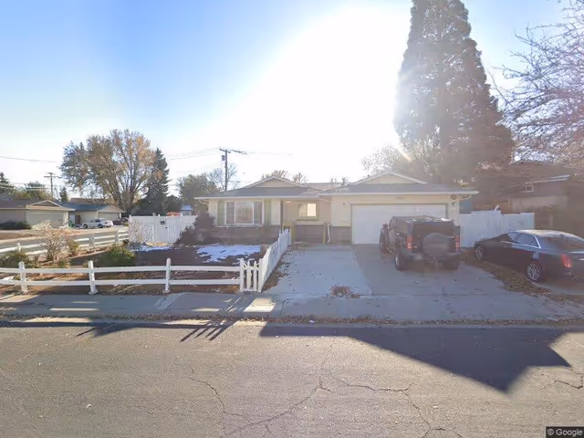 Single-story residential house with a white picket fence, a driveway with two parked cars, and some trees in the background under a bright sky.