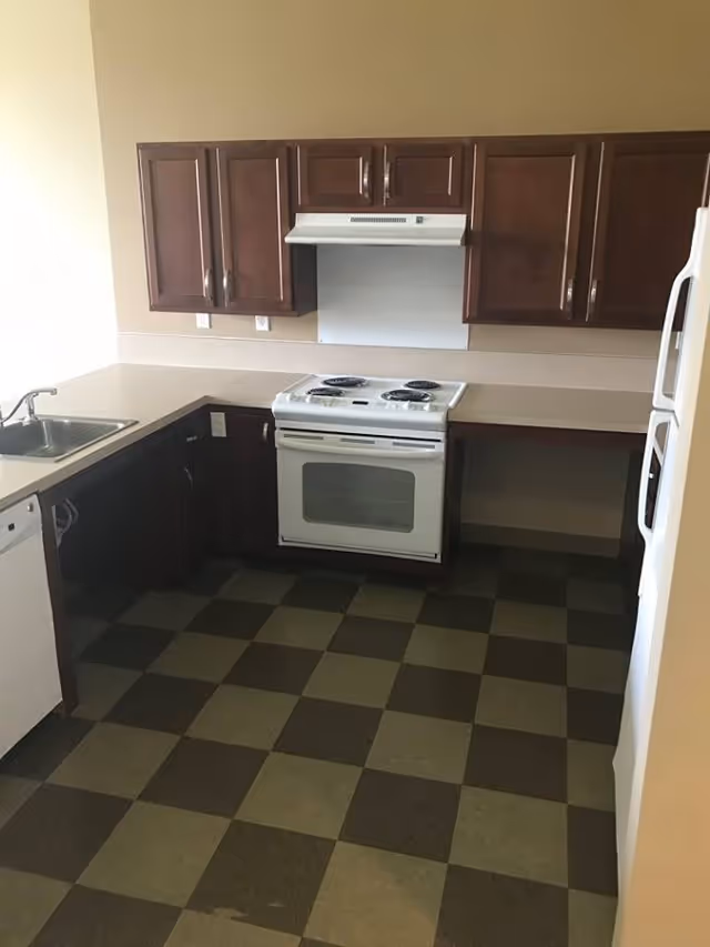 A kitchen with dark brown upper and lower cabinets, a white electric stove with four burners, a white refrigerator, a white dishwasher, a stainless steel sink, and a checkered floor with alternating dark and light brown tiles.