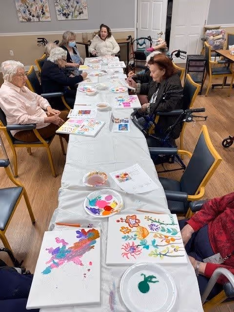 A group of elderly individuals seated around a long table engaged in a painting activity. The table is covered with a white cloth and has various paint palettes, brushes, and colorful paintings in progress. The room has wooden flooring and light-colored walls with framed artwork.