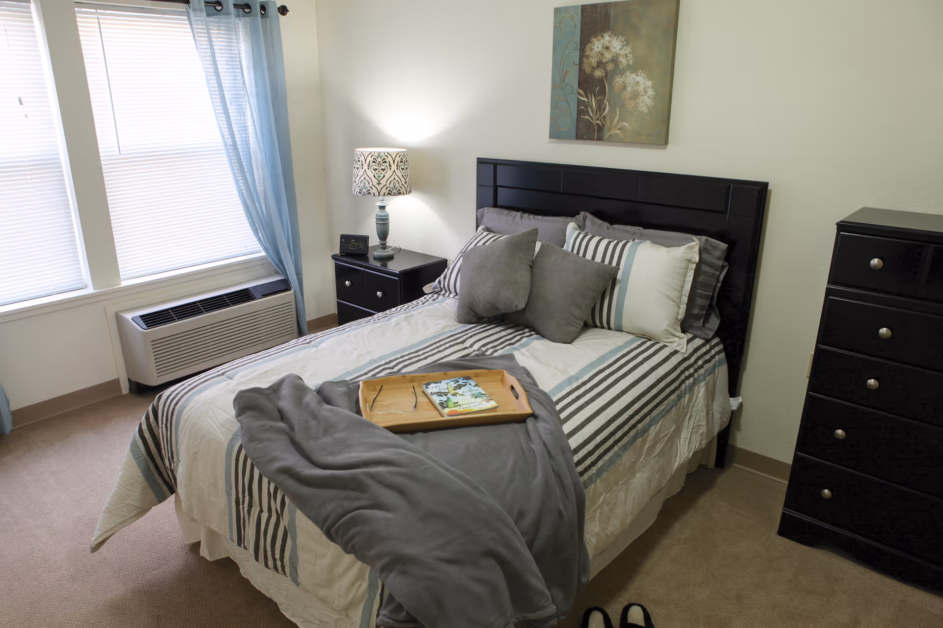 A neatly made bed with striped bedding and multiple pillows in a bedroom. A wooden tray with a book and a pair of glasses rests on the bed. Next to the bed is a nightstand with a decorative lamp and a digital clock. A window with blue curtains and blinds is on the left side, and a black dresser with drawers is on the right side. A floral painting hangs on the wall above the bed.