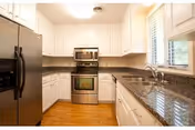 Bright U-shaped kitchen with stainless steel refrigerator and oven, white cabinets, granite countertops, hardwood floors, and a window over the sink.