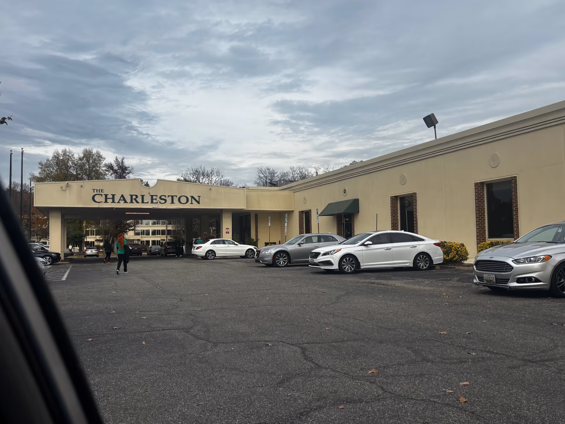Exterior view of The Charleston Senior Community building with a covered entrance and several parked cars in front. People are walking near the entrance under a cloudy sky.