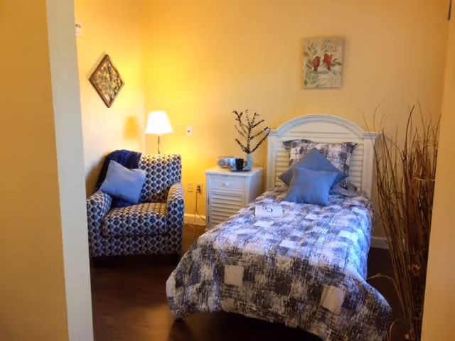 A cozy bedroom with a single bed covered in a blue and white patterned quilt and two blue pillows. Next to the bed is a white nightstand with a small lamp, a radio, and a decorative branch. To the left, there is a patterned armchair with a blue cushion. The walls are painted yellow, and there is a framed artwork on the wall above the chair. A bundle of tall dried branches is placed on the right side of the bed.