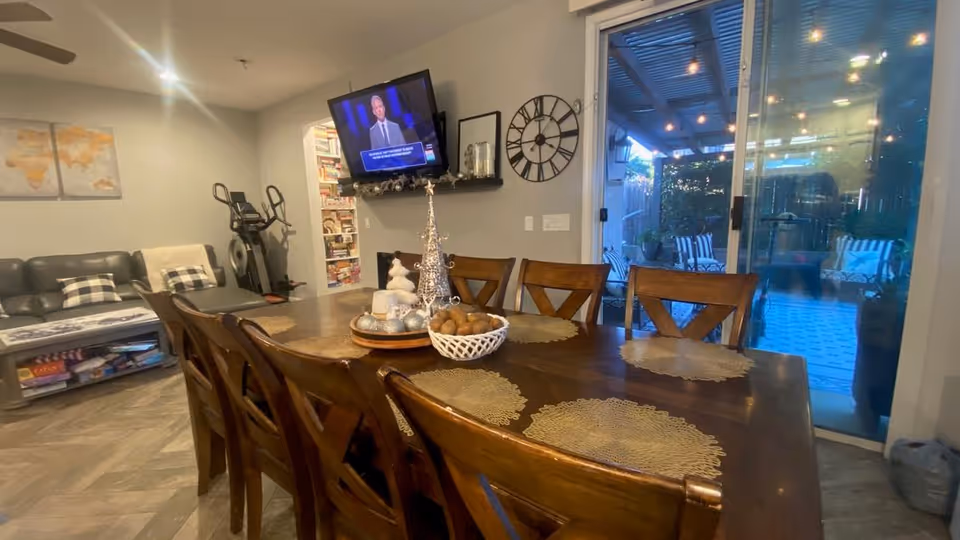 A wooden dining table with placemats and a decorative centerpiece in an open-plan living area with a TV, couch, and sliding glass door to a patio.