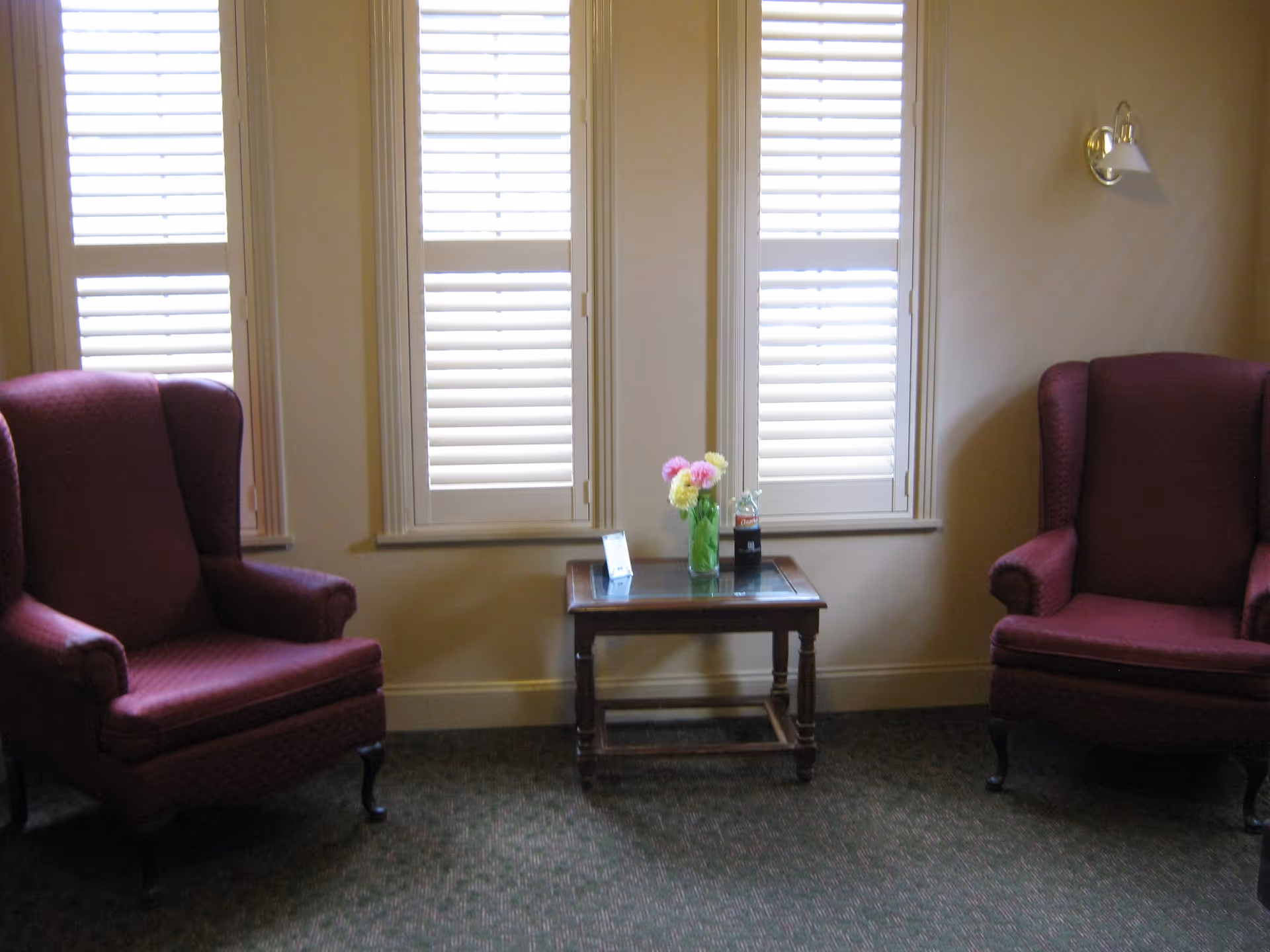 A cozy sitting area with two maroon wingback chairs facing each other, separated by a small wooden table with a glass top. On the table, there is a vase with colorful flowers and a small decorative item. Behind the table are three tall windows with white plantation shutters, and a wall-mounted light fixture is visible on the right side.