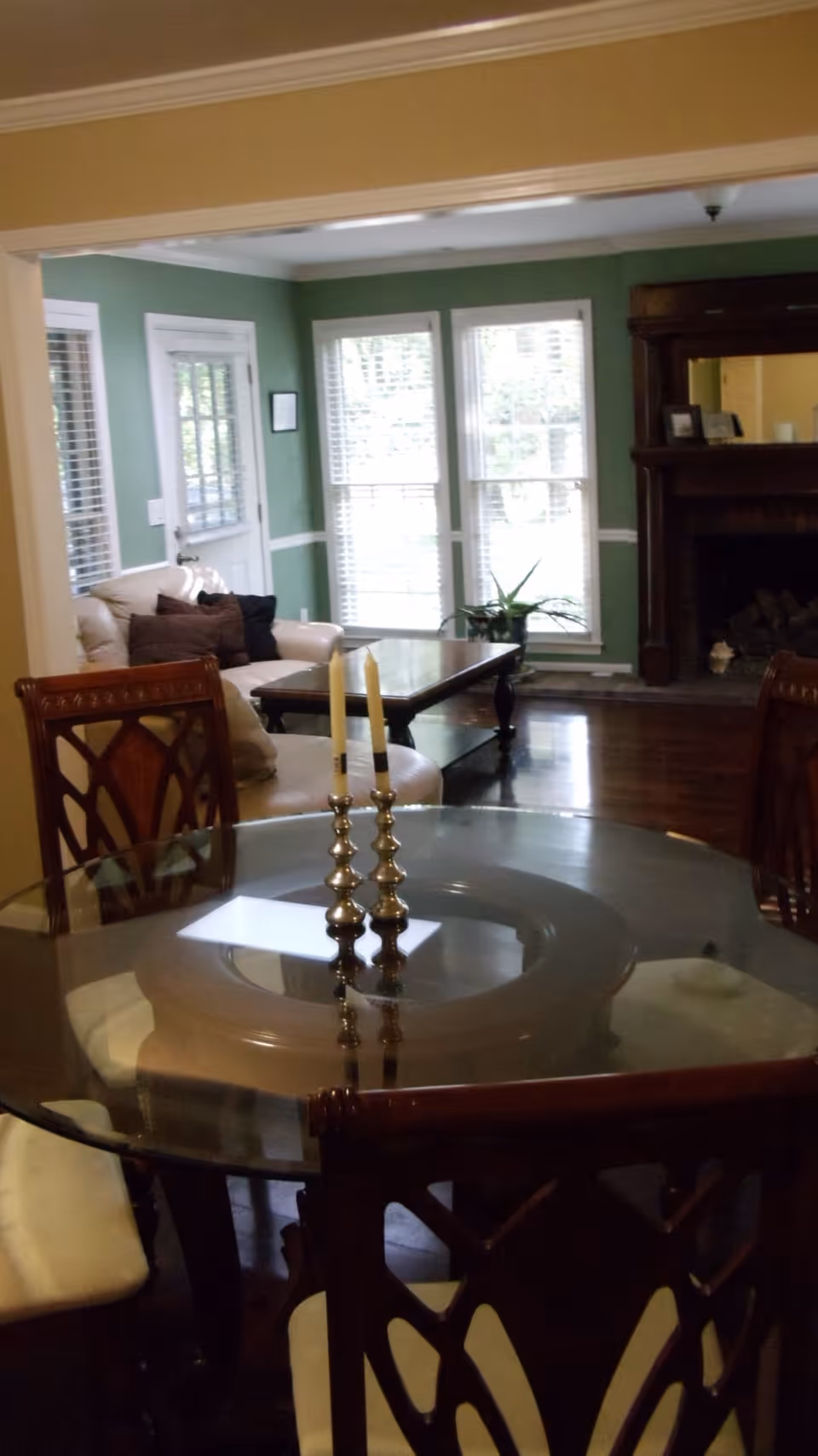 Glass-top dining table with candlesticks in the foreground and a connected living room with sofa, coffee table, large windows, and a fireplace beyond.