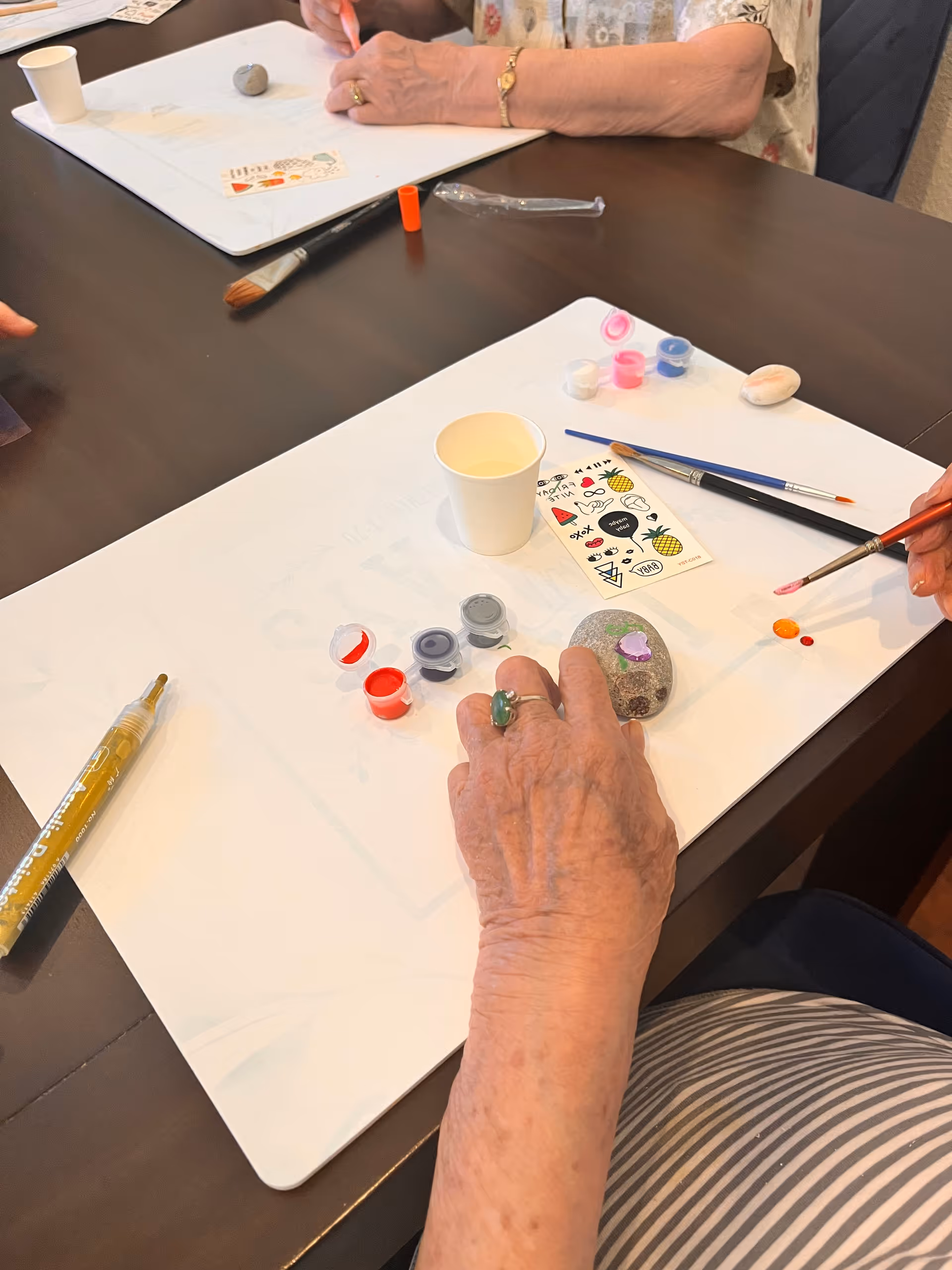 Two elderly individuals seated at a table engaged in a rock painting activity. One person is holding a paintbrush applying paint to a rock, while the other person's hands rest on the table near painting supplies including small paint containers, brushes, a paper cup, and decorative stickers.