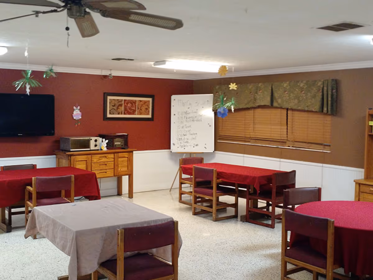 A senior living facility common room with several tables covered with red and beige tablecloths, wooden chairs, a ceiling fan, a wall-mounted TV, a wooden cabinet with a microwave and a radio, a whiteboard with a schedule, and windows with green valances.