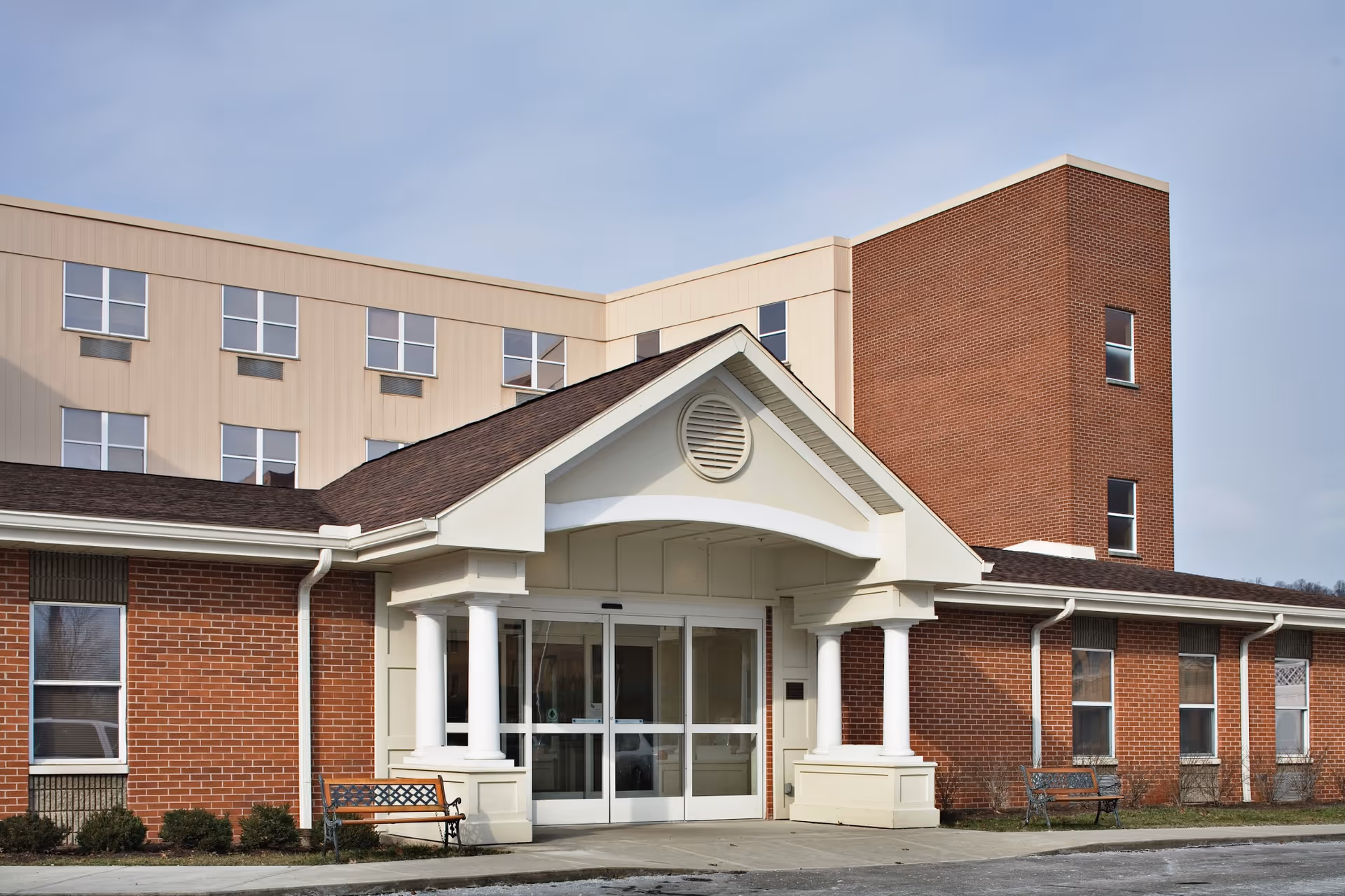 Exterior view of a senior living facility building with a brick facade and beige siding. The entrance features a covered porch with white columns and glass double doors. There are benches on either side of the entrance and multiple windows on the building. The sky is clear with some clouds.