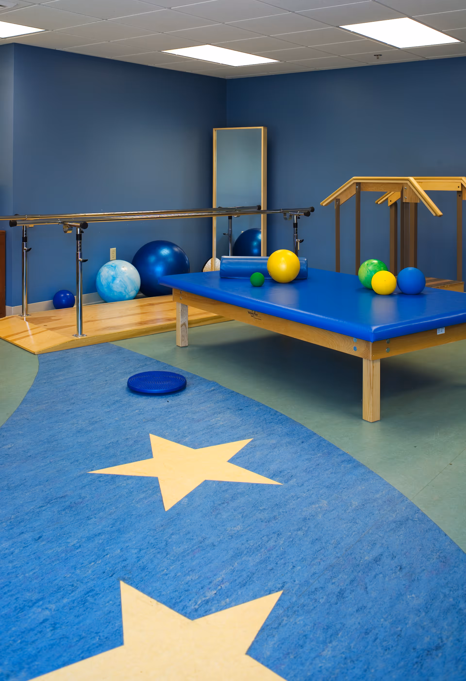 Physical therapy room with a blue padded therapy table holding various colored therapy balls, parallel bars for walking exercises, a wooden staircase with handrails, a full-length mirror, and a blue floor mat with yellow star designs.