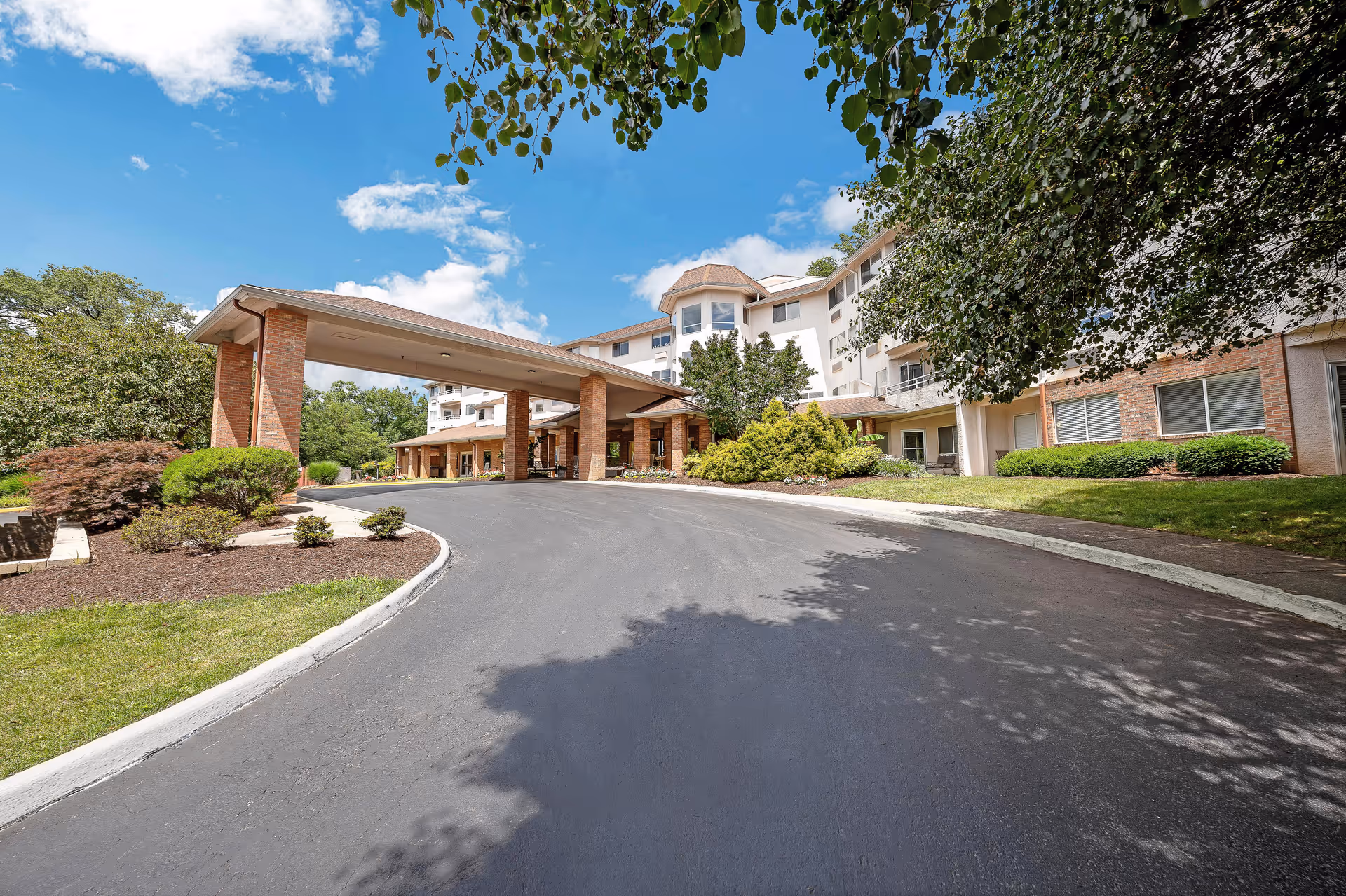 Front exterior view of Holiday Elm Park Estates, showing a multi-story building with a covered entrance driveway, surrounded by landscaped greenery and trees under a blue sky with some clouds.