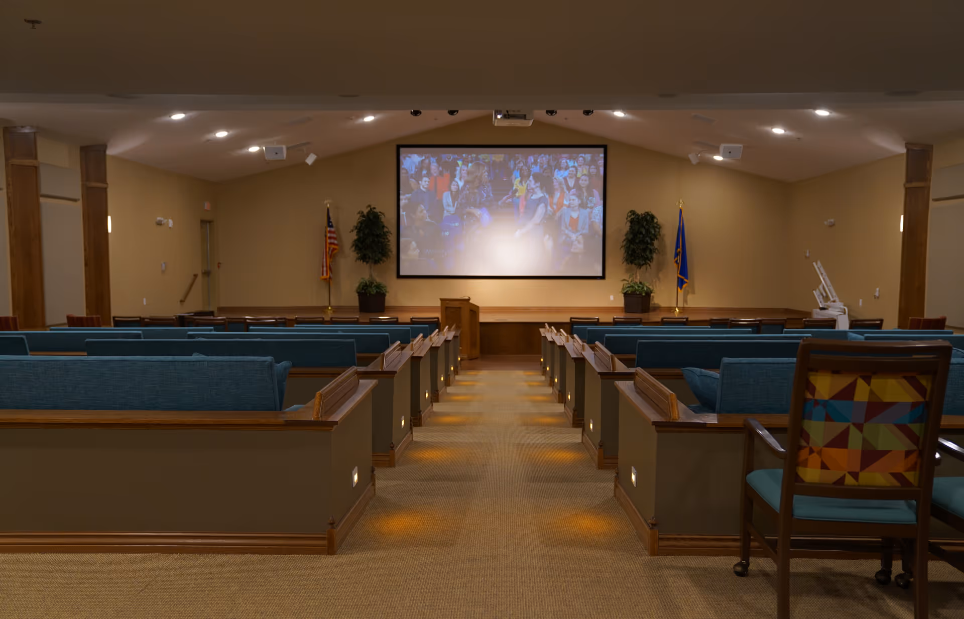 Interior auditorium-style room with rows of blue upholstered benches facing a large projection screen and podium at the front.
