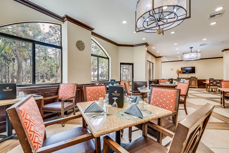 Dining room with tables set for a meal, red-patterned chairs, large arched windows, and chandeliers.