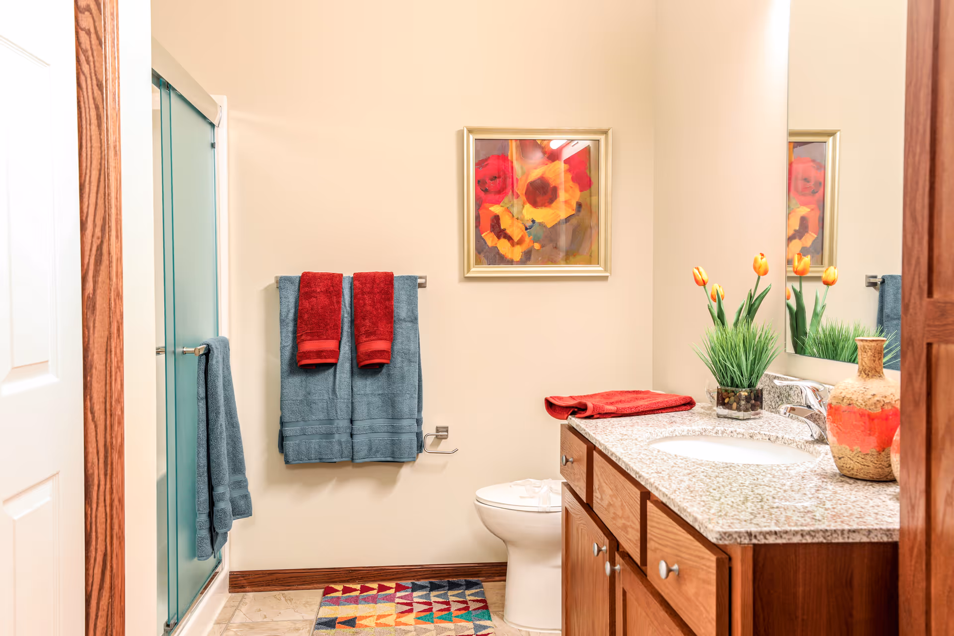 A clean and well-lit bathroom featuring a granite countertop with a sink, wooden cabinets, a toilet, and a glass-enclosed shower. The wall has a colorful framed painting of flowers, and there are blue and red towels hanging on towel racks. A small plant with orange tulips and a decorative vase are placed on the countertop. A multicolored geometric patterned rug is on the floor.